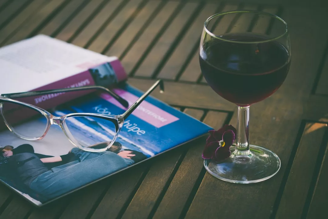 A glass of red wine, eyeglasses, and an open magazine rest on a wooden table, radiating a sense of luxury lifestyle, with a small purple flower beside the glass.