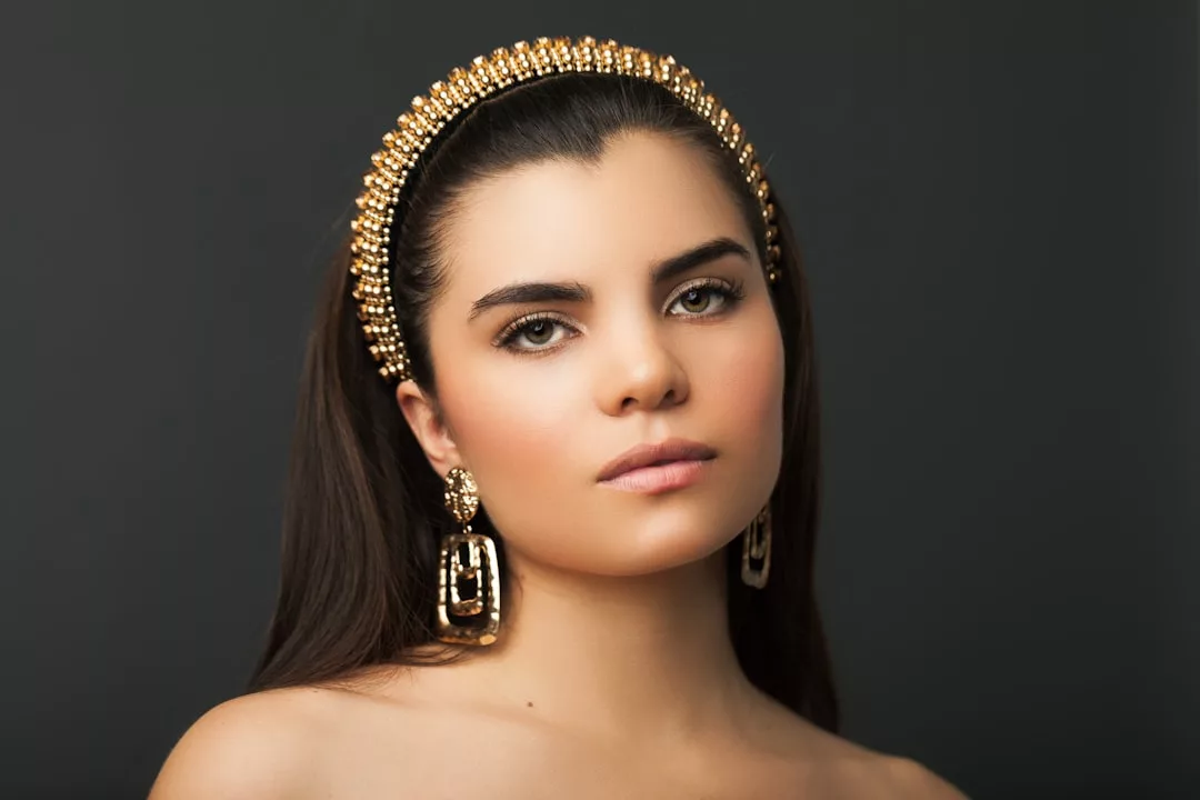 Woman with long dark hair wearing a gold beaded headband and large gold earrings, posing against a plain dark background, embodies timeless luxury and modern fashion.
