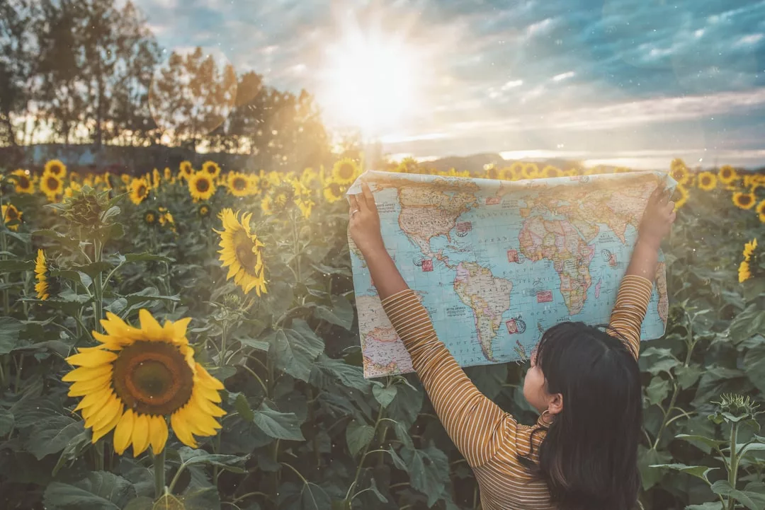 A person stands in a sunflower field at sunset, holding up a world map and facing away from the camera, embodying Glamesia’s sense of wanderlust and fashion.