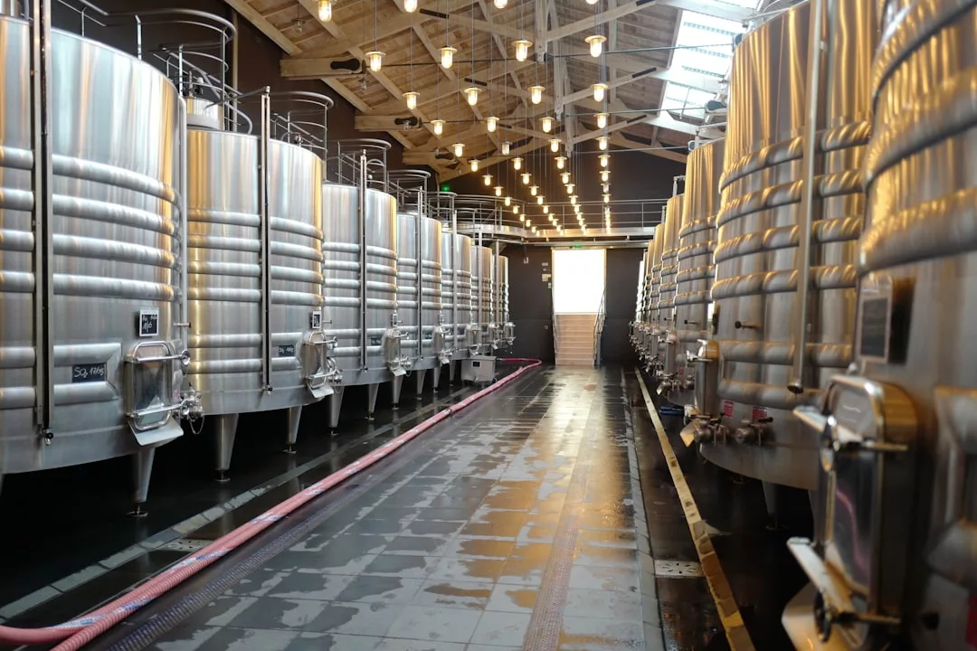 A row of large stainless steel fermentation tanks lines both sides of a brightly lit industrial facility with a clean concrete floor, reflecting a modern lifestyle dedicated to quality and precision.