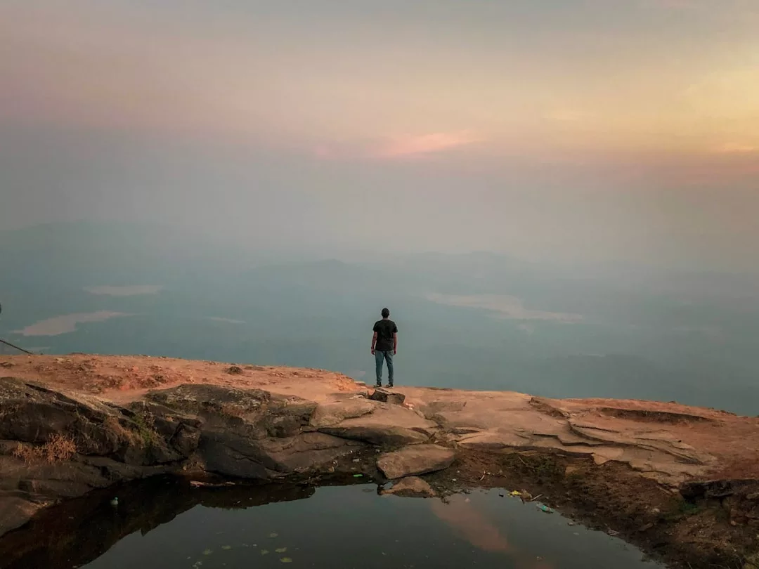 A person stands near the edge of a rocky cliff overlooking a misty valley at sunset, exuding a sense of fashion and grace, with a small pool of water in the foreground reflecting their stylish presence.