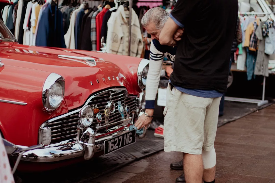 Two people examine the front bumper and license plate of a vintage red car at an outdoor market, where racks of clothing highlight a unique blend of fashion and classic style in an open-air lifestyle setting.