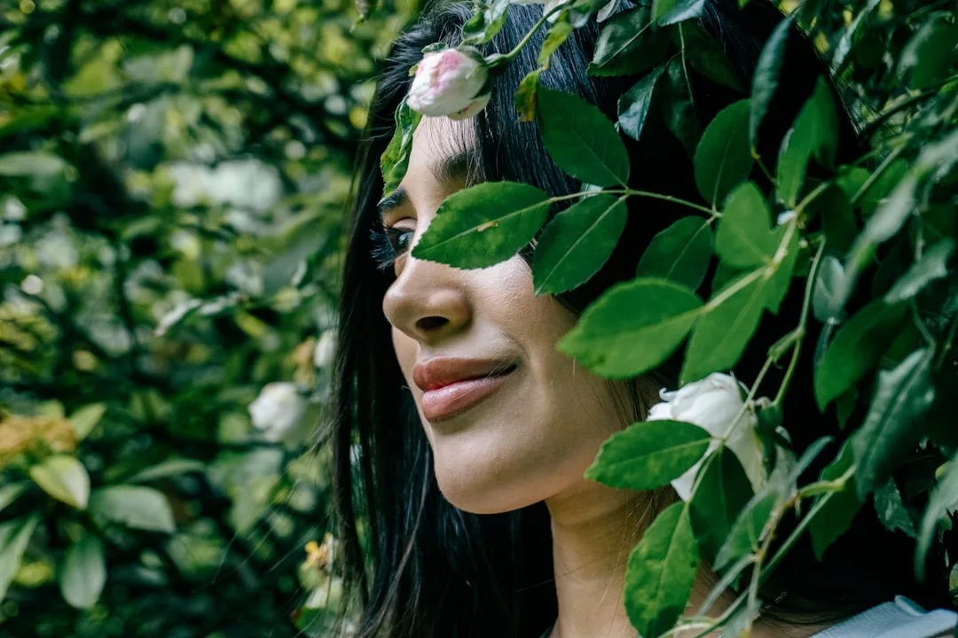 A woman stands among green leaves and white roses in a Glamesia outdoor setting, with foliage partially covering her face.
