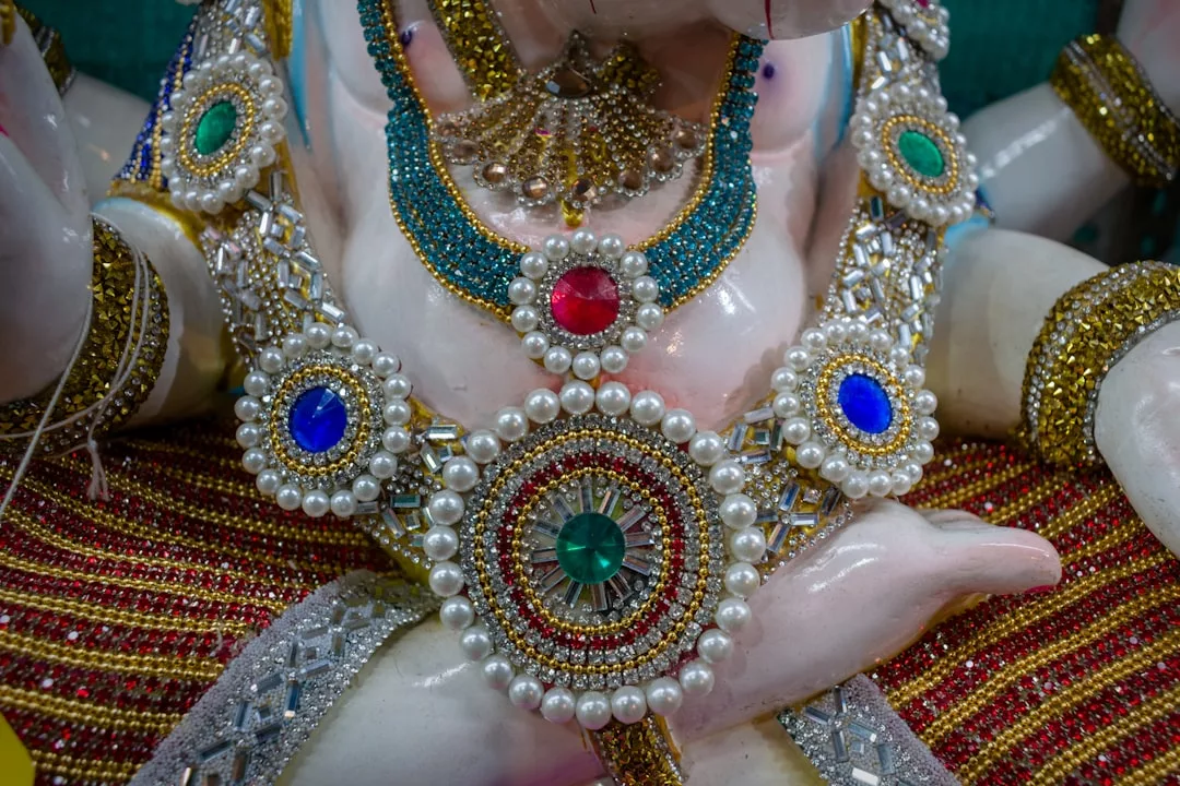 Close-up of a decorative statue adorned with elaborate luxury jewelry, featuring beads, pearls, and colorful gemstones in red, blue, and green.