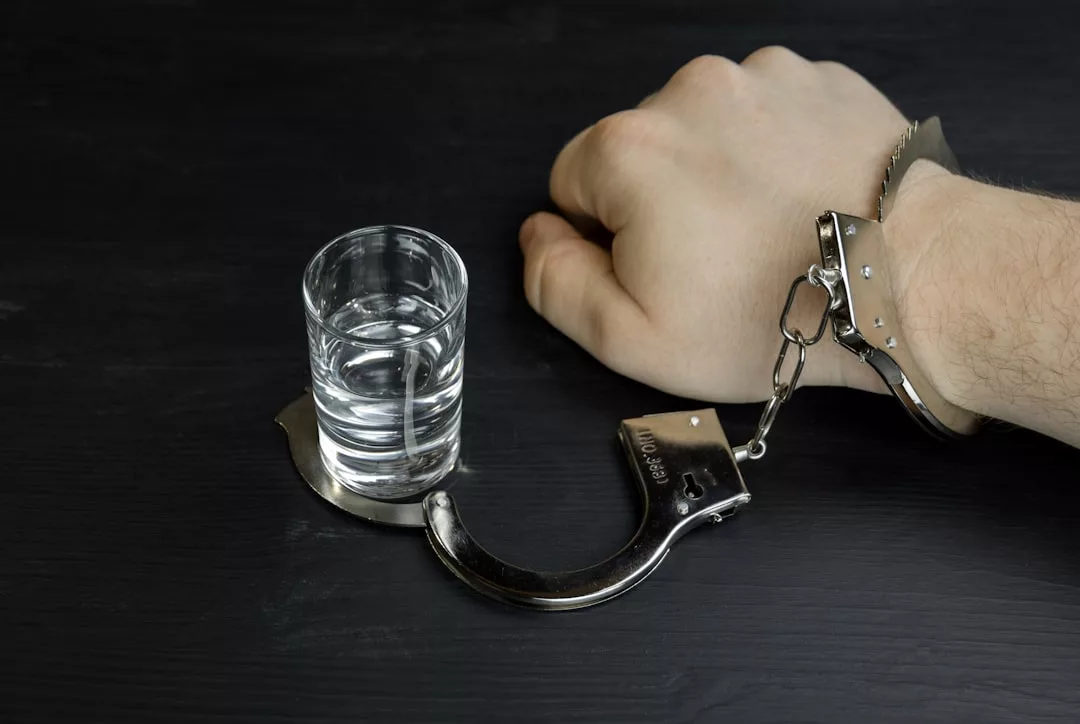 A person's wrist is handcuffed to a shot glass filled with clear liquid, both resting on a dark surface—an edgy nod to lifestyle and fashion rebellion.