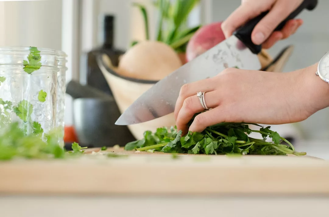 A person chops fresh cilantro on a wooden cutting board with a large kitchen knife, showcasing culinary fashion next to a glass jar and bowl of vegetables.