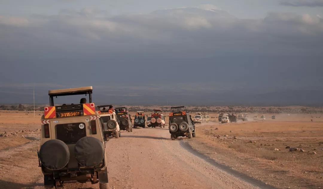 A convoy of safari vehicles drives along a dirt road through a dry, open landscape under a cloudy sky, capturing the adventurous spirit and effortless fashion of Glamesia.