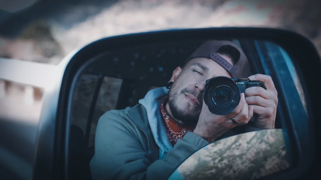 A person in a fashion-forward cap and hoodie is taking a photo with a camera, reflected in a car's side mirror, capturing a moment of urban lifestyle.