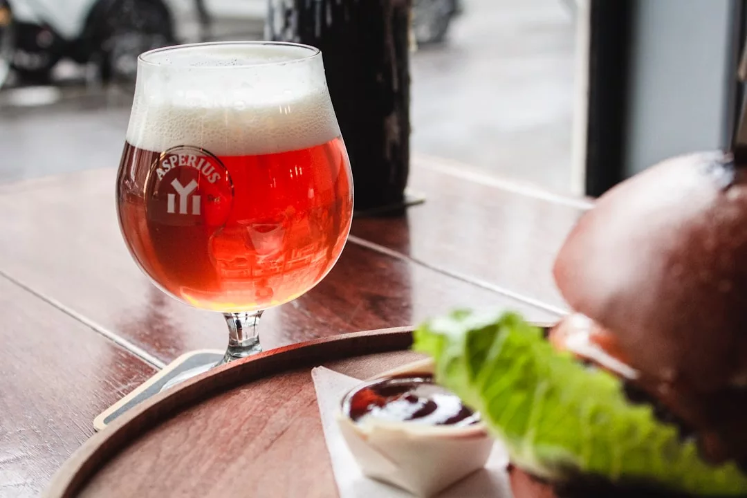 A glass of amber beer sits on a wooden table next to a plate with a burger, lettuce, and a small cup of sauce, capturing the essence of a laid-back lifestyle.