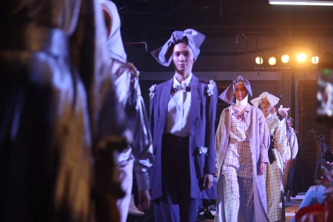 Models walk down the runway in luxurious, elaborate outfits with large headpieces, illuminated by bright stage lights at Glamesia’s indoor fashion show—a true celebration of high-end lifestyle and glamour.
