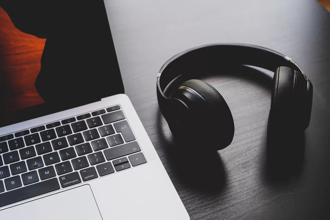 A pair of black over-ear headphones, blending seamlessly with modern lifestyle, is placed next to a laptop on a dark wooden surface.