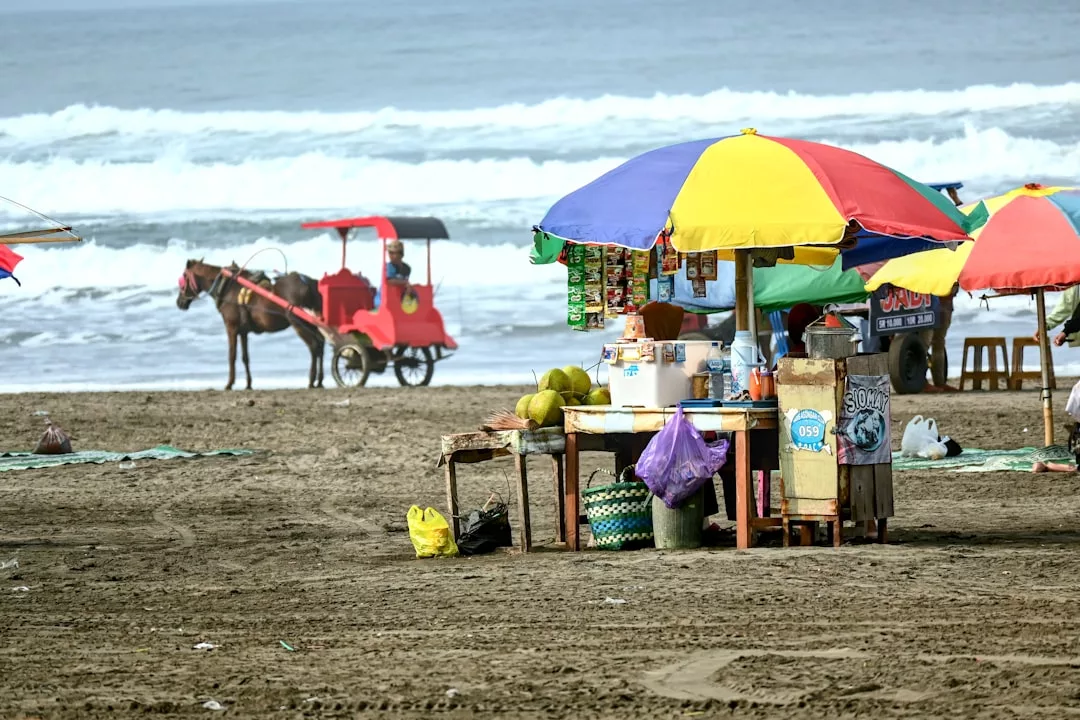 A colorful beachside food stall with umbrellas stands on the sand, offering a glimpse of luxury living as a horse-drawn carriage passes by and ocean waves roll in the background.
