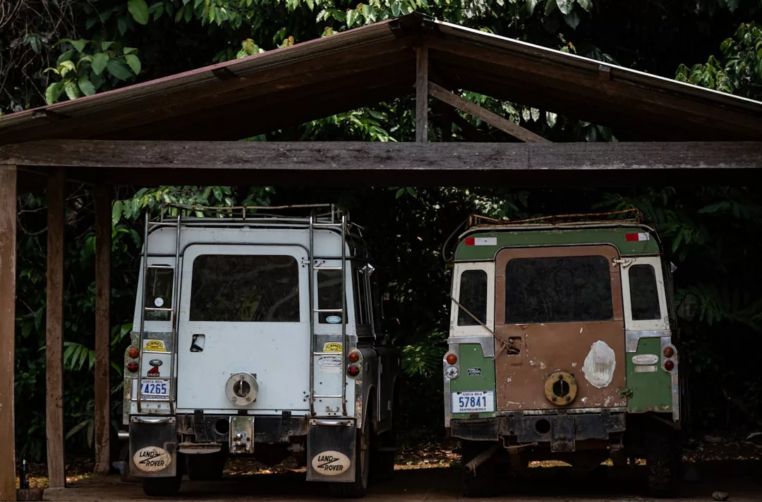 Two old Land Rover vehicles, one white and one brown, are parked side by side under a wooden carport with green foliage in the background, reflecting a timeless lifestyle and understated luxury.