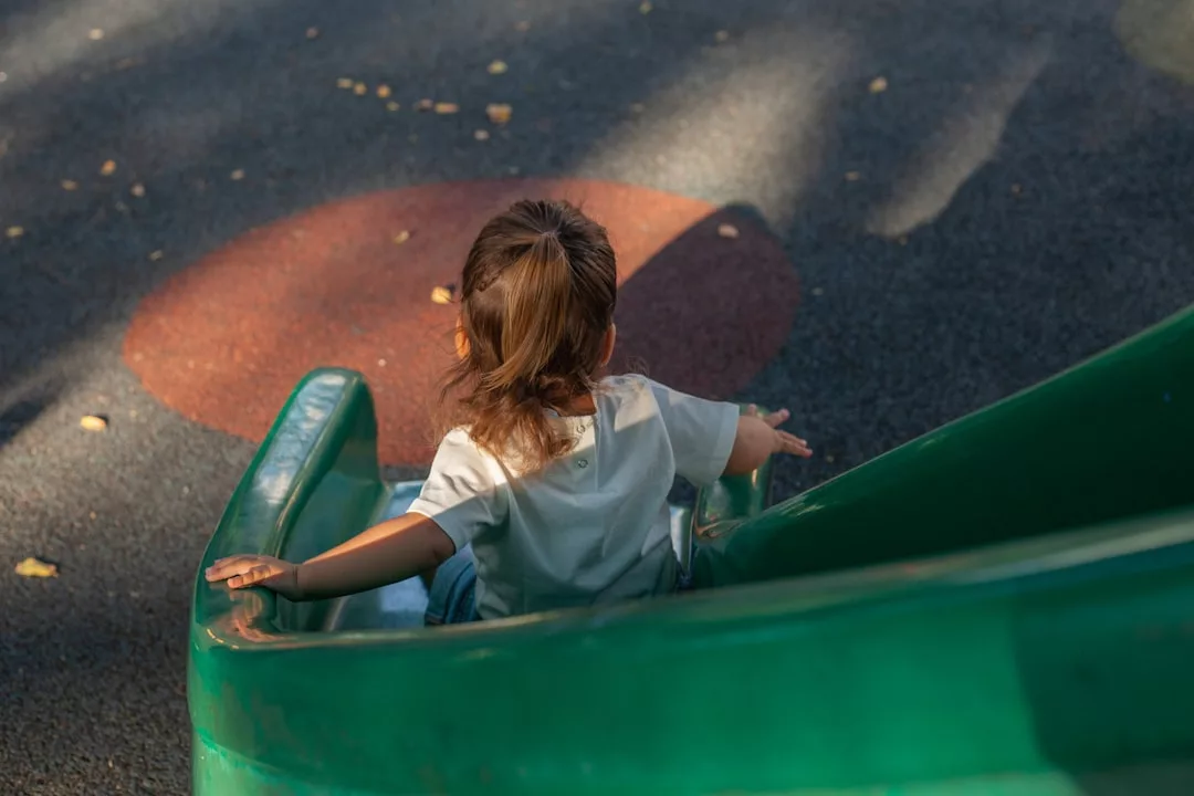 A young child with brown hair, seen from behind, sits at the top of a green playground slide under daylight, embodying casual fashion with effortless charm.