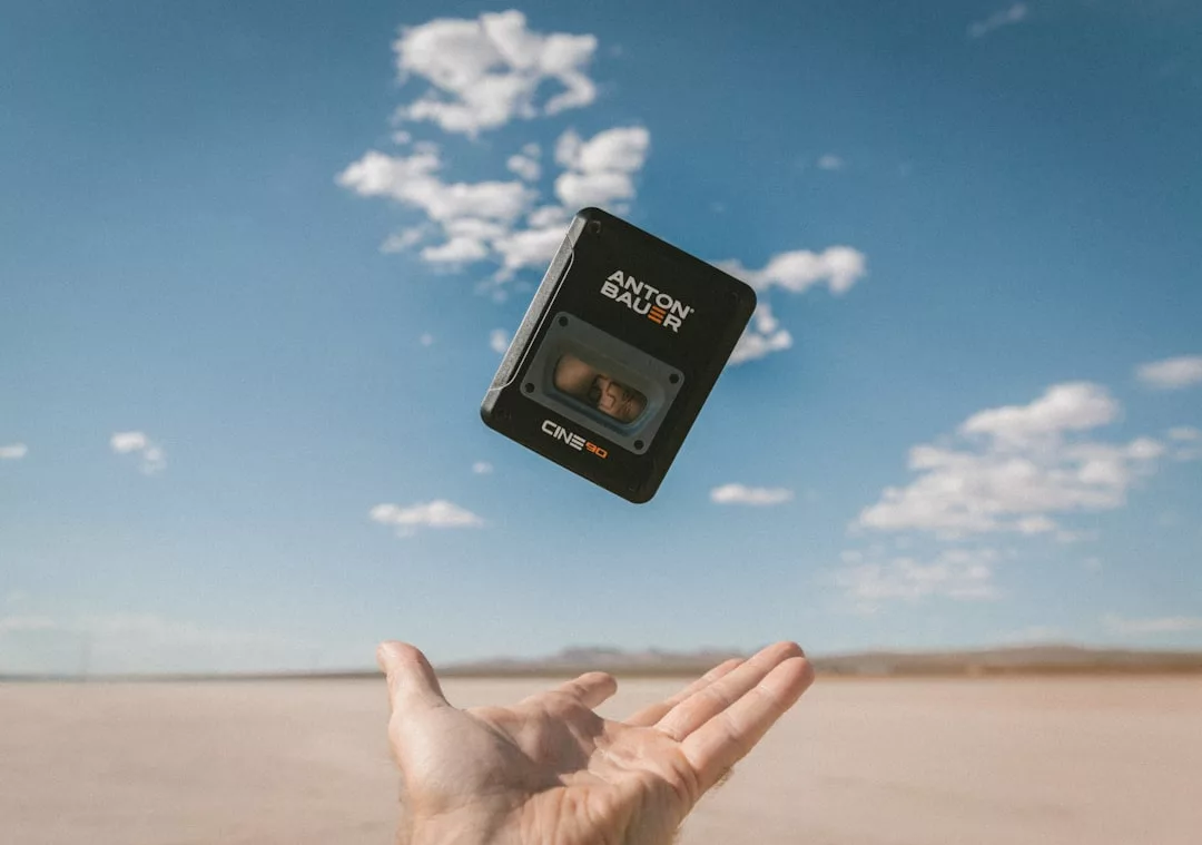 A hand tosses an Anton Bauer Cine 150 battery into the air against a backdrop of blue sky and scattered clouds, capturing a moment of luxury amidst the rugged desert landscape.