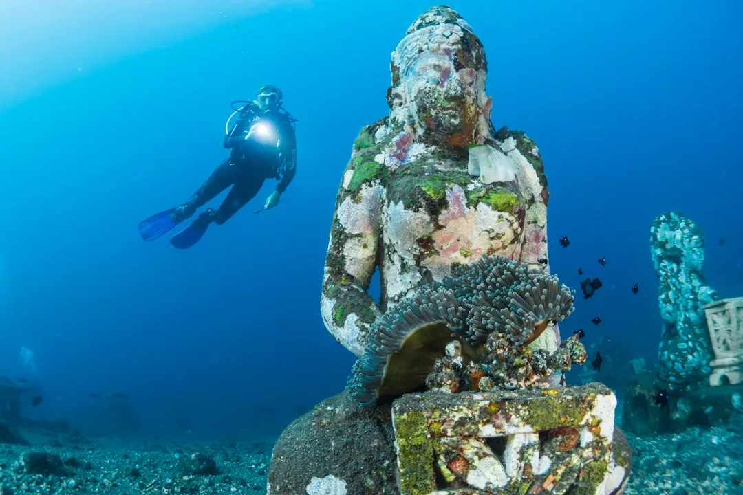 A scuba diver enjoys a luxury lifestyle as they swim near a coral-encrusted underwater statue on the ocean floor, surrounded by blue water and vibrant marine life.