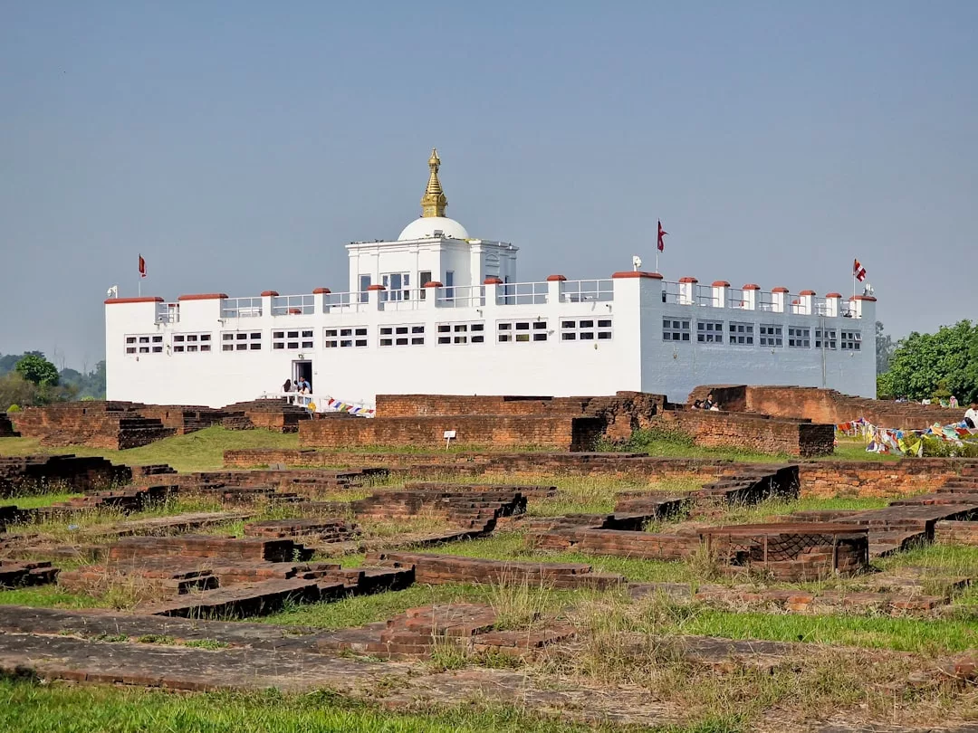 A white rectangular building with a golden spire, known as the Maya Devi Temple, stands behind ancient brick ruins under a clear sky, evoking a sense of timeless luxury and serene lifestyle.