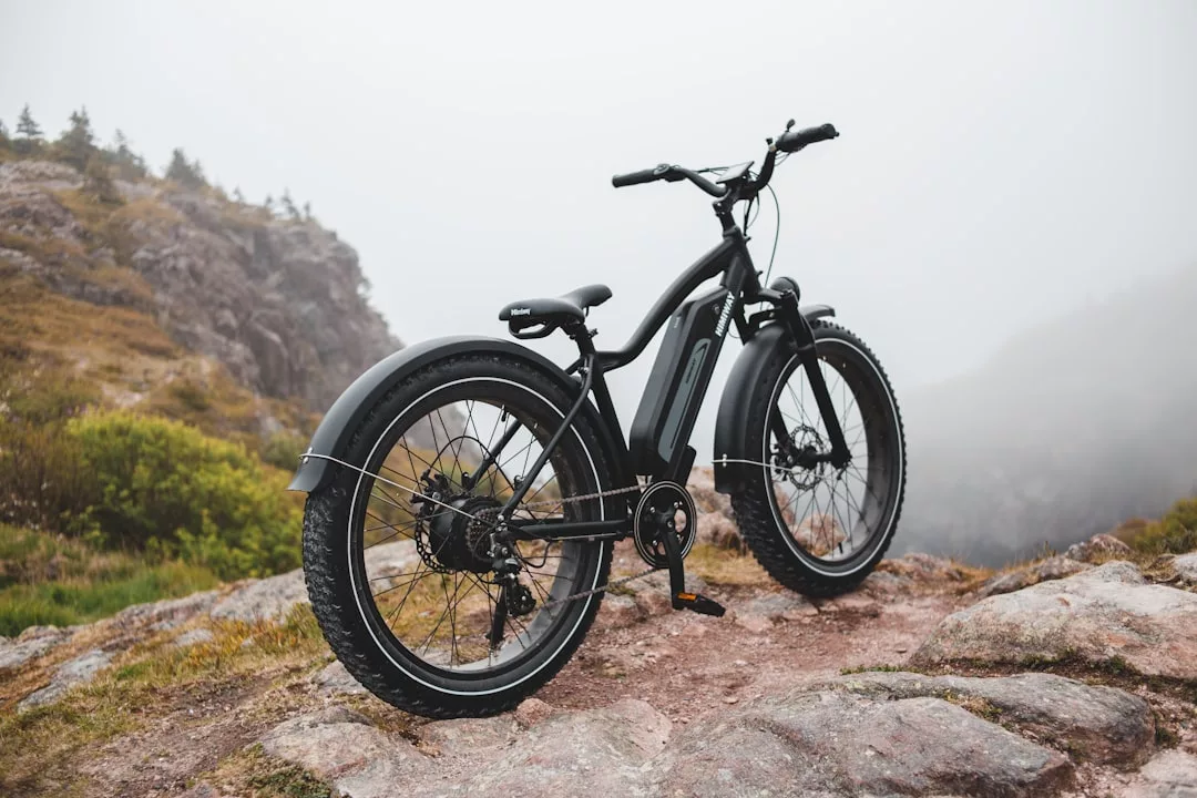 A black electric mountain bike, blending luxury with adventure, is parked on a rocky trail with misty hills and lush greenery in the background.