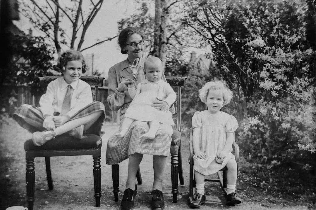 Black and white photo of an older woman sitting outdoors with three young children, two beside her and one baby on her lap, surrounded by trees and plants, radiating timeless Glamesia style amid natural luxury.