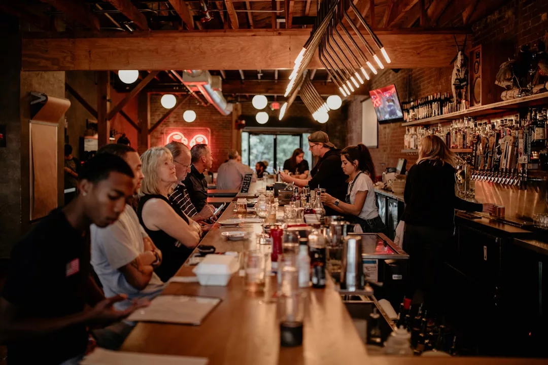 People sit and socialize at a busy, fashion-forward bar with wooden interiors; some face the bartender while others use their phones. Shelves of bottles and hanging lights add to the Glamesia charm.