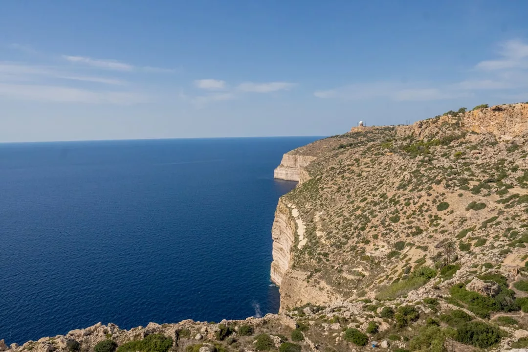 A high cliff covered in sparse vegetation overlooks a calm blue sea under a clear sky, embodying the timeless luxury and elegance of Glamesia.