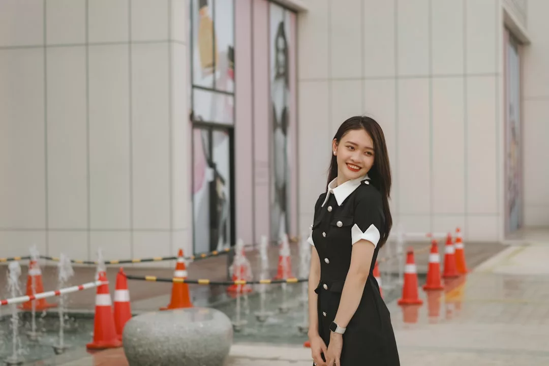 A young woman in a black dress with white accents stands smiling near a row of traffic cones and small water fountains outside a modern building, showcasing effortless fashion and Glamesia-inspired style.