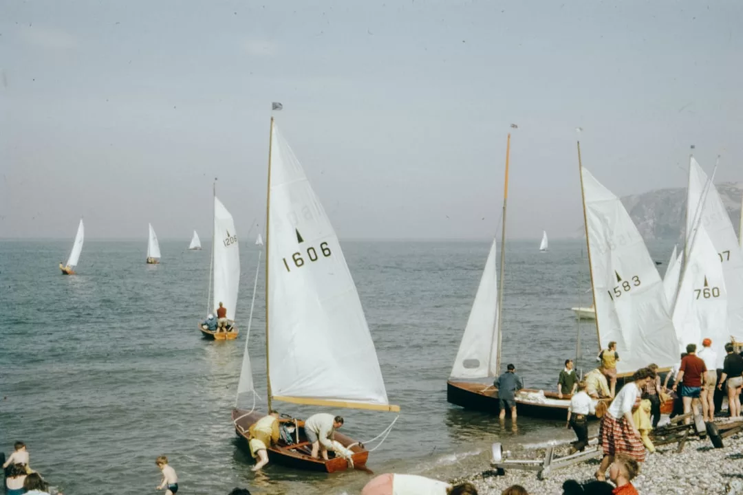 Several small sailboats with white sails gather near a pebbled shore, where people prepare to board or launch them into the calm sea under a clear sky, embracing a touch of luxury by the water’s edge.