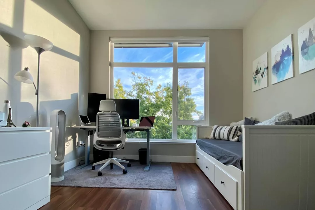 A modern home office with a desk, ergonomic chair, monitors, and a window with a view of trees. A daybed and artwork add style on the right, while a white dresser enhances the sophisticated lifestyle on the left.