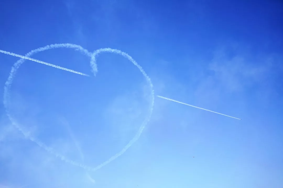 A jet leaves a white contrail in the sky, intersecting with a large heart shape formed by a previous smoke trail against a blue sky—a glimpse of Glamesia lifestyle painted above.