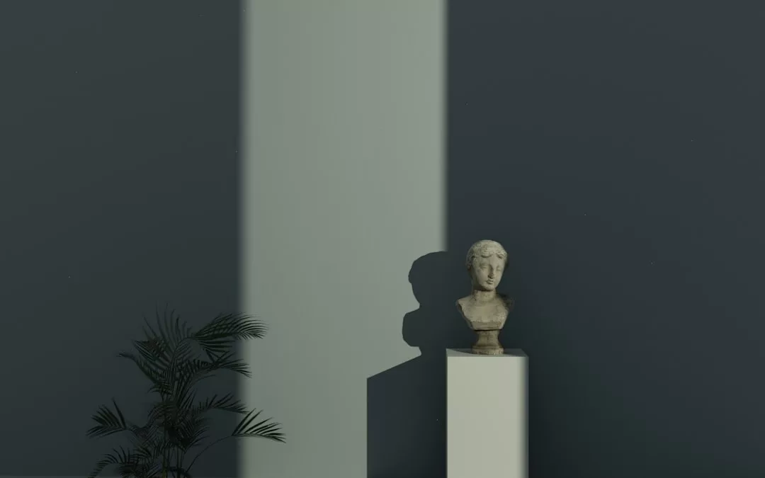 A classical bust on a pedestal is illuminated by a vertical beam of light against a dark wall, with a potted plant in the lower left corner, creating a refined lifestyle ambiance.