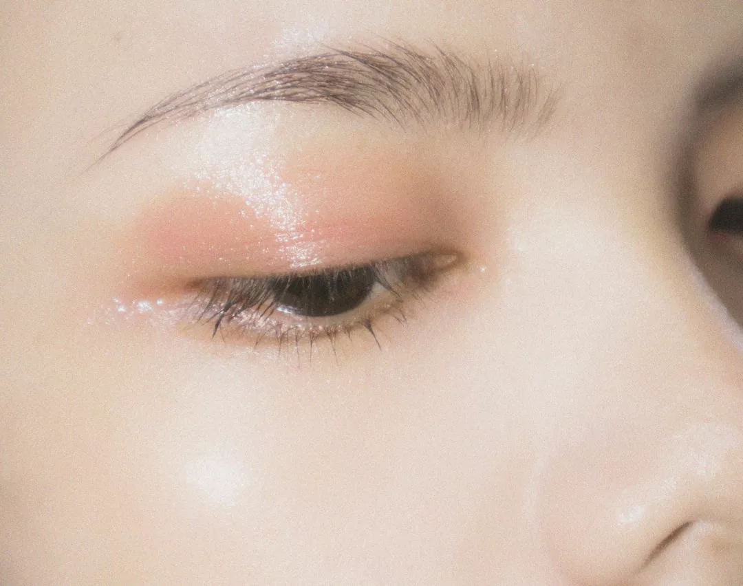 Close-up of a person's closed eye with glossy pink eyeshadow and a well-groomed eyebrow, capturing smooth, glowing skin—a true moment of Glamesia luxury in fashion beauty.
