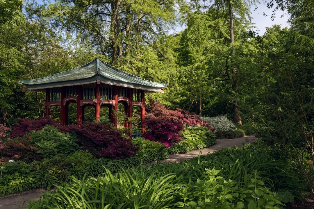 A luxury red and green gazebo stands surrounded by lush trees, shrubs, and flowering plants along a winding garden path.