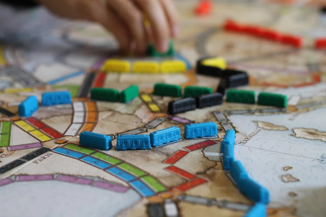 Close-up of a board game in progress, showing colorful plastic train pieces placed on a map, with a hand reaching towards the pieces—a stylish touch to any modern lifestyle.
