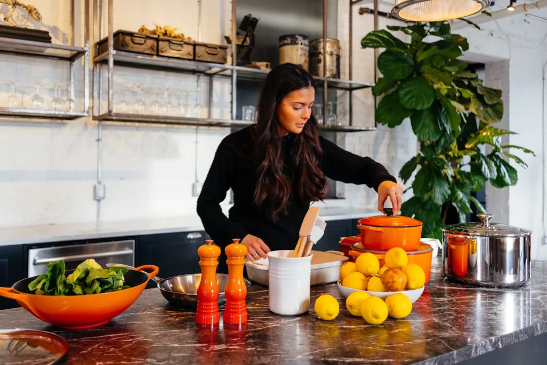 A woman in a kitchen prepares food at a counter with pots, bowls, lemons, spinach, and Glamesia kitchen utensils, bringing a touch of fashion and flair to her cooking routine.