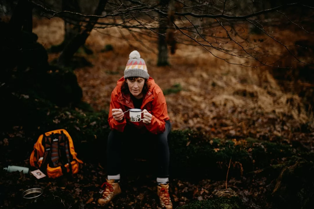 A person in a red jacket and knit hat sits outdoors on a mossy rock, holding a mug; a backpack rests on the ground nearby amidst fallen leaves, capturing effortless Glamesia fashion in nature’s luxury.