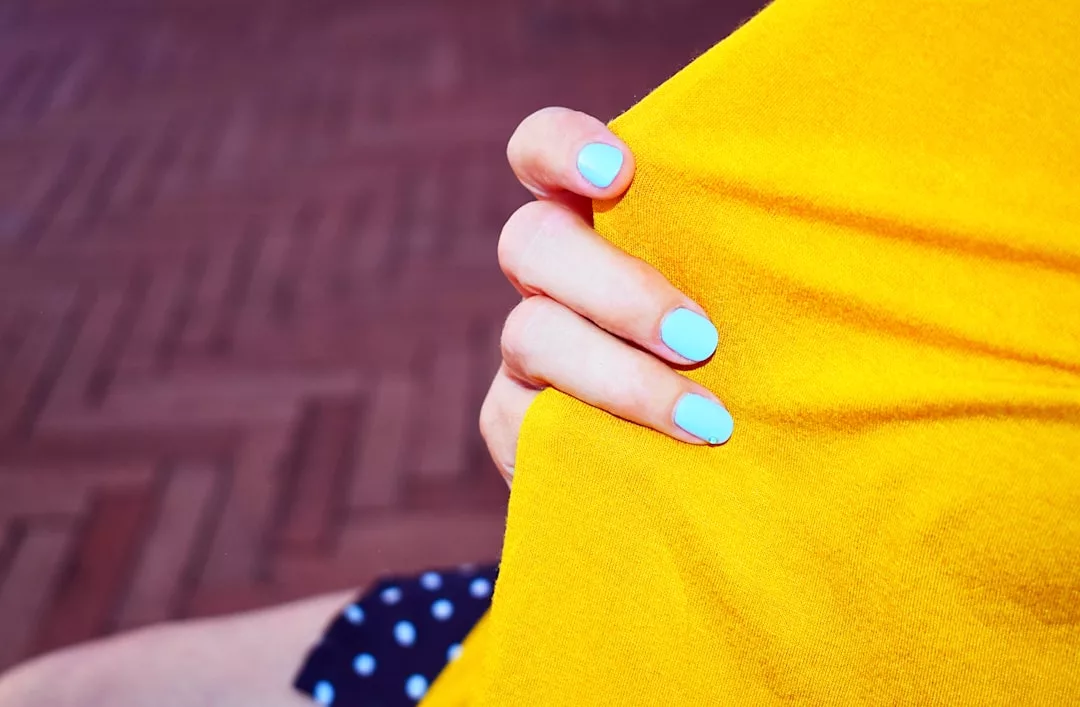 A hand with light blue nail polish holds a yellow fabric, capturing a moment of fashion and lifestyle against a brick-patterned ground and dark fabric with white dots in the background.
