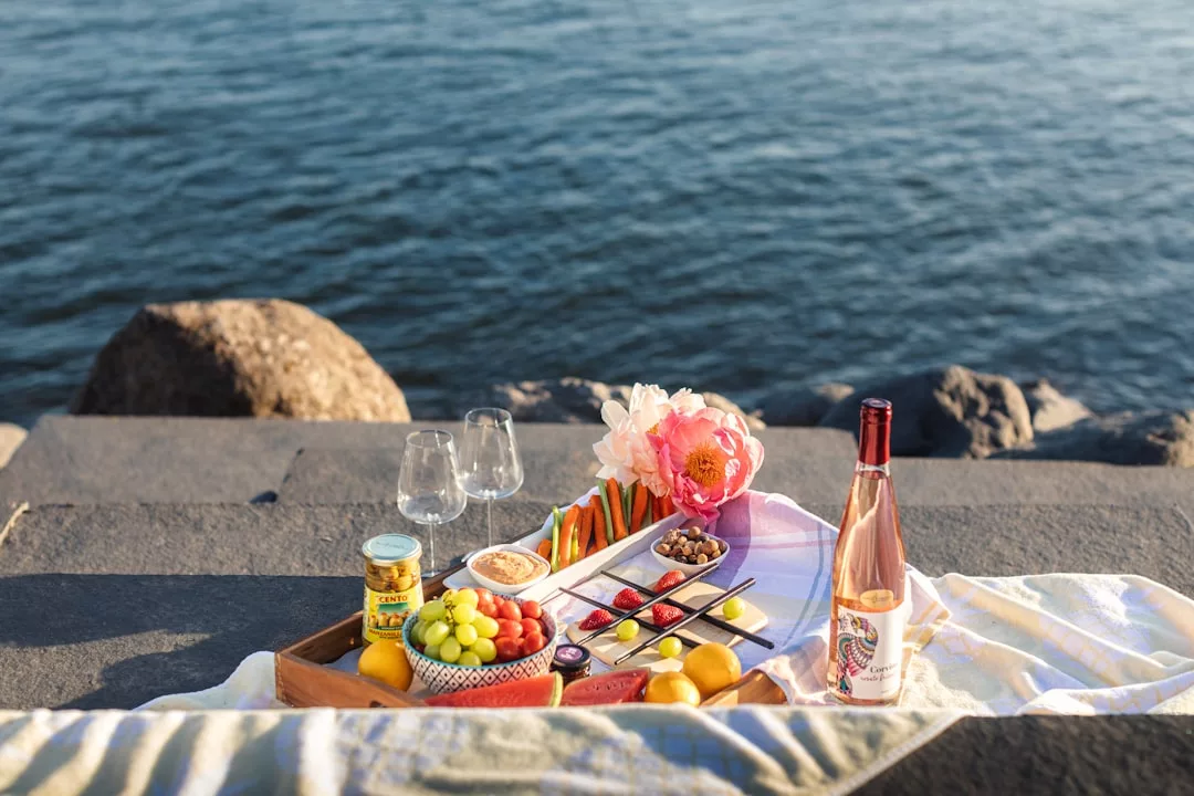 A Glamesia-inspired picnic setup on a stone surface by the water, featuring a tray of fruits, snacks, a bottle of rosé, two glasses, and a bouquet of flowers for a touch of luxury lifestyle.