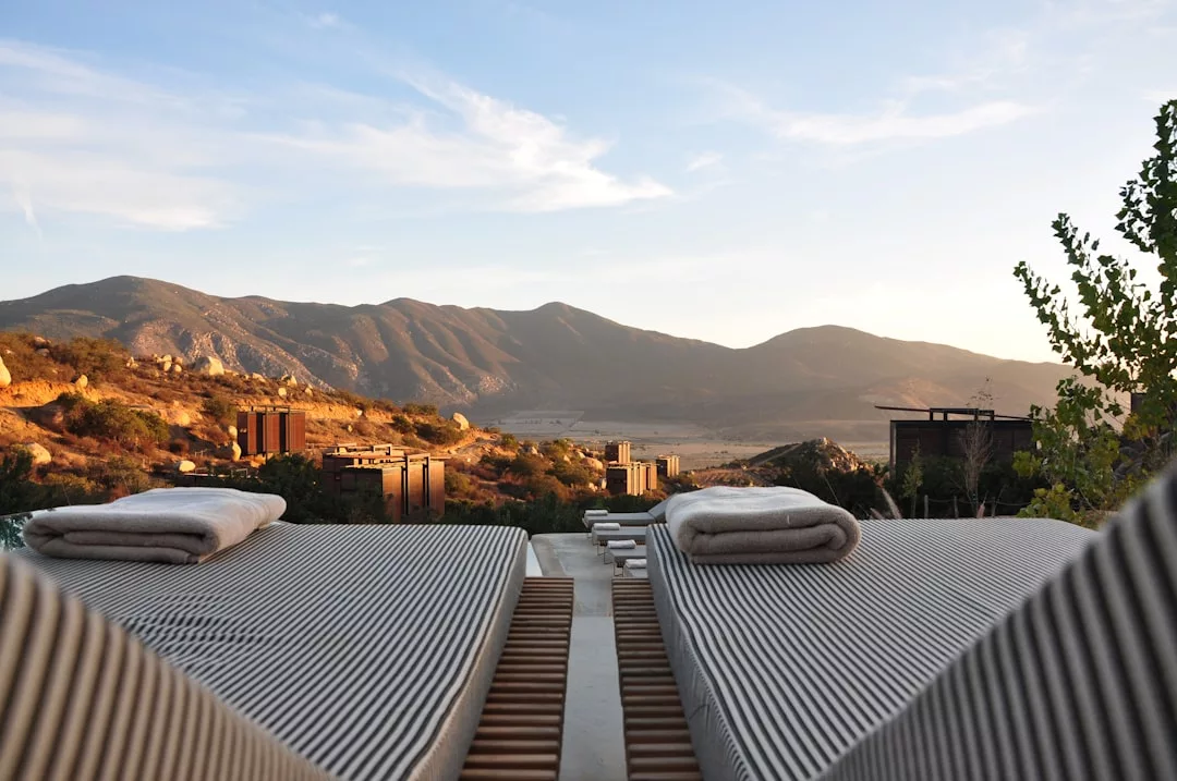 Two luxury lounge chairs with towels face a scenic mountain landscape at sunset, with stylish buildings scattered on the hillside in the background.