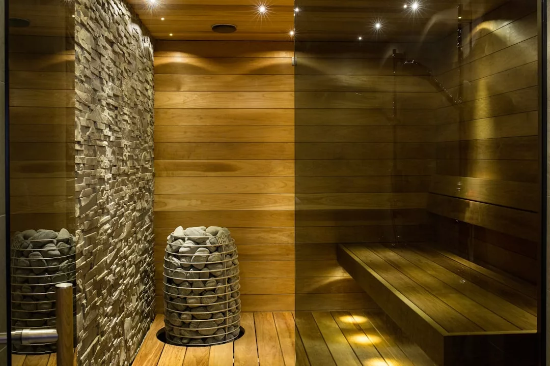 Modern wooden sauna room with a stone accent wall, a metal basket filled with sauna rocks, and built-in wooden bench seating under warm lighting for an elevated luxury lifestyle experience.