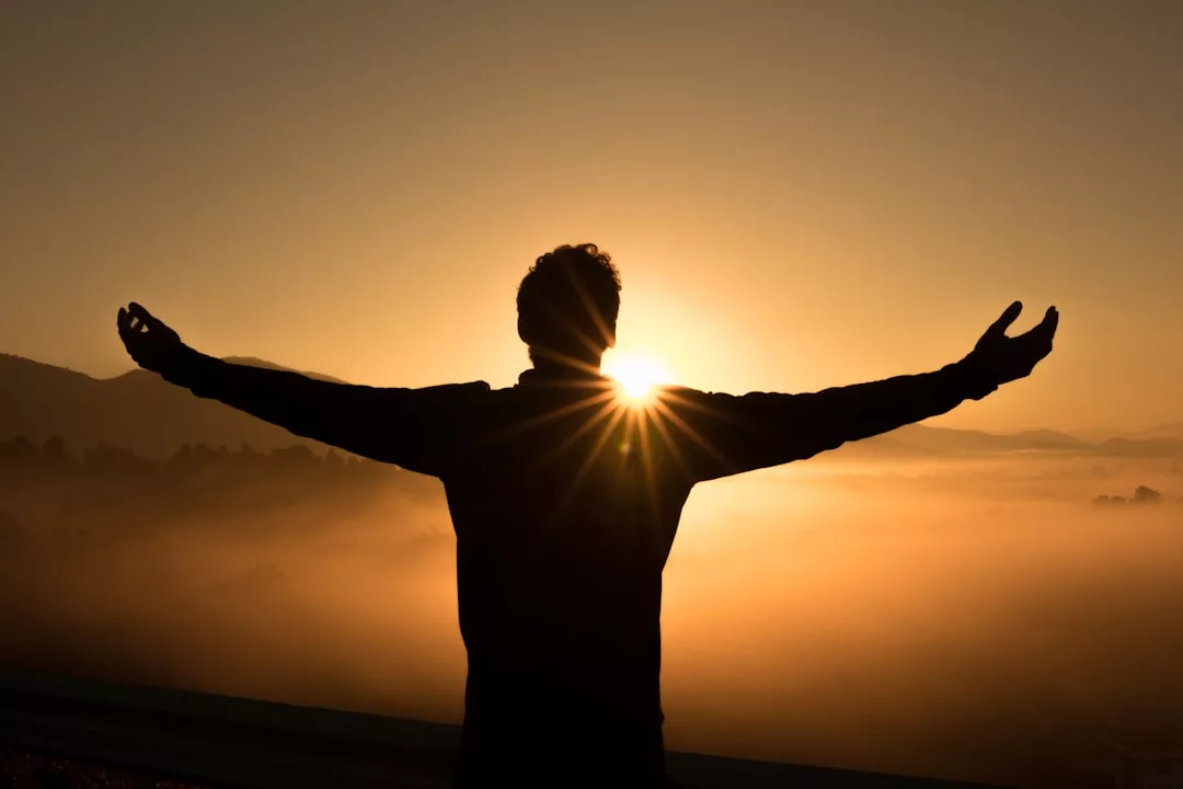A person stands with arms outstretched facing the sunrise, silhouetted against a misty landscape, embodying serene fashion and the spirit of Glamesia.
