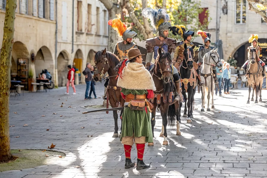 People in historical costumes, some on horseback and one standing, gather in a cobblestone plaza with onlookers and stone buildings, evoking a glimpse into the luxury lifestyle of a bygone era.