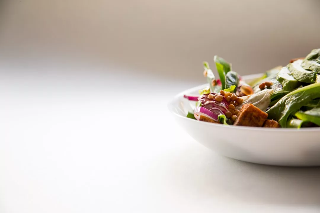 A close-up of a white bowl filled with salad, featuring greens, sliced red onions, nuts, and croutons, epitomizing luxury lifestyle dining on a plain light background.