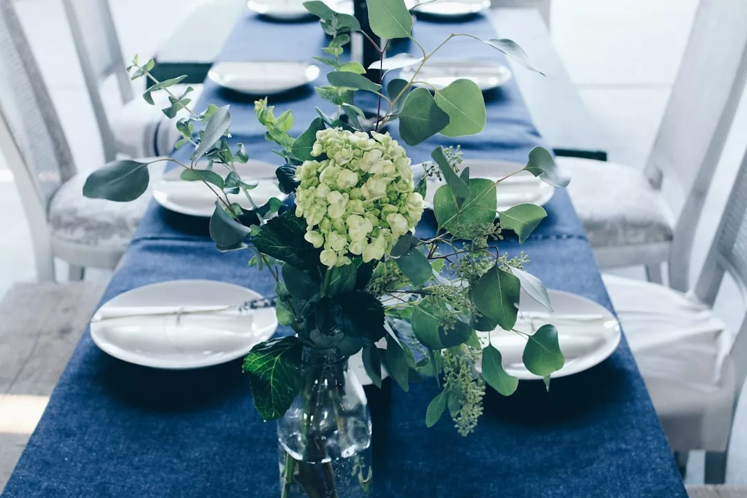 A glass vase with green and white flowers serves as a fashion-forward centerpiece on a dining table set with white plates and blue table runners.