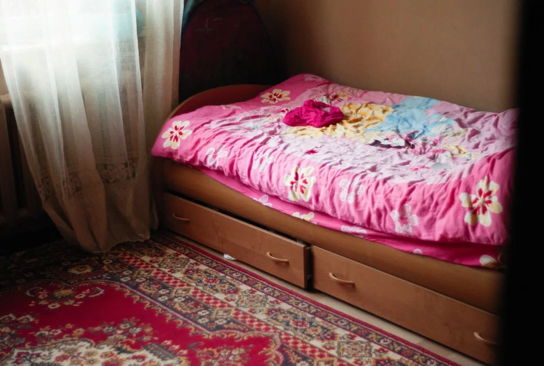 A small bed with a pink floral blanket and some fashion-forward clothes on top, next to a window with sheer curtains and a patterned carpet on the floor.