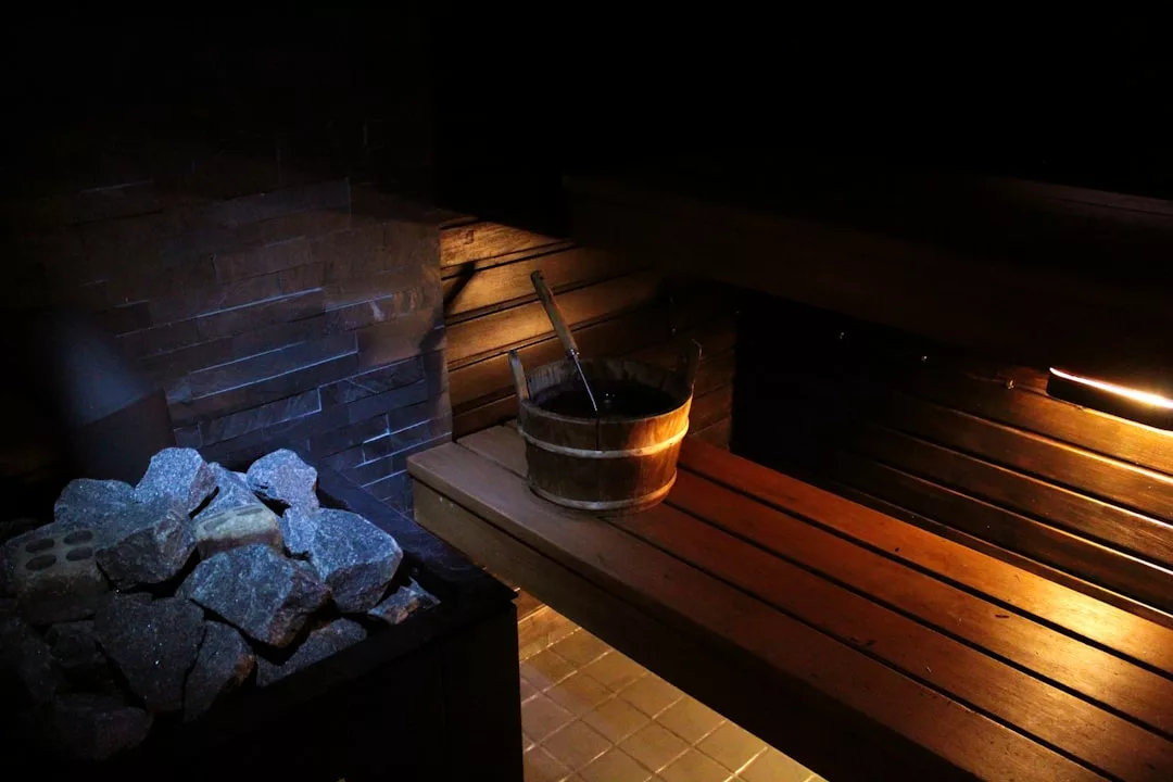A wooden sauna interior with a bucket and ladle on a bench, dim lighting, and a pile of sauna stones on a heater in the corner, offering a touch of Glamesia luxury to your wellness lifestyle.