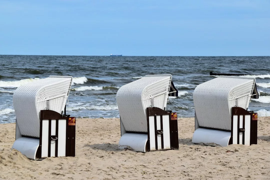 Three white wicker beach chairs sit on the sandy shore, facing the sea with gentle waves and a distant ship under a partly cloudy sky—capturing the essence of luxury lifestyle by Glamesia.
