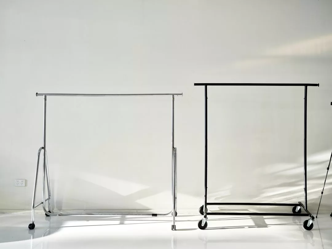Two empty rolling garment racks, one silver and one black, stand side by side against a plain white wall in a brightly lit room, reflecting a modern Laxary lifestyle.