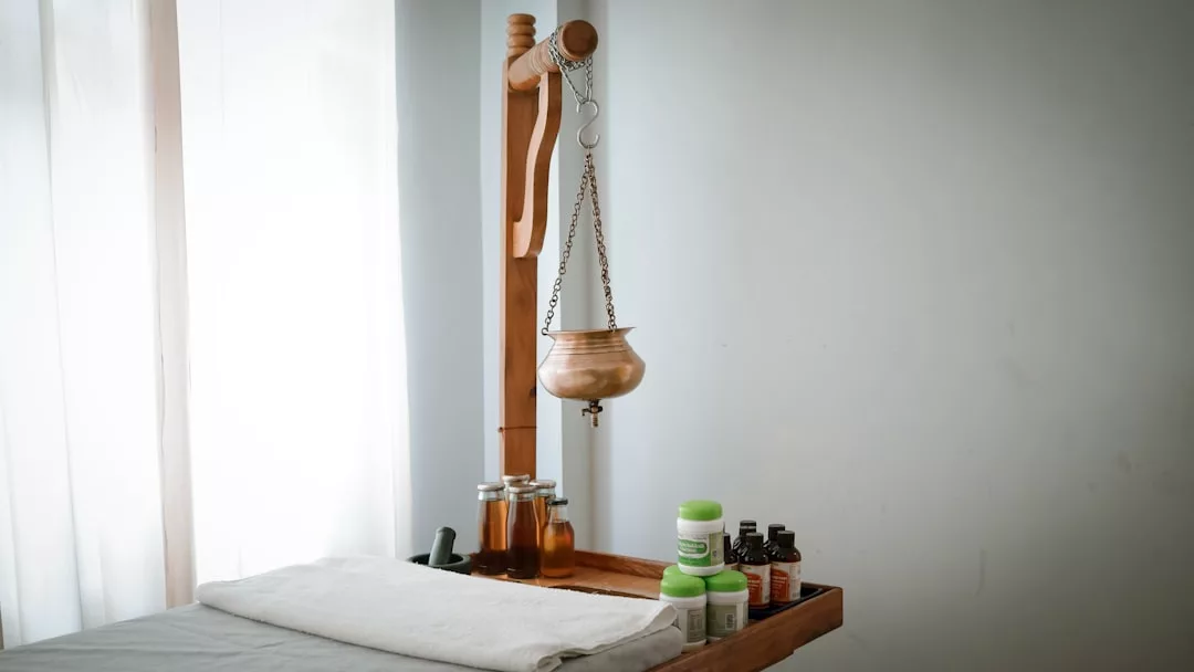 A luxury massage table with a folded towel, herbal oil bottles, supplement containers, and a hanging brass oil pot creates an inviting atmosphere in this spa or wellness room—perfect for an elevated lifestyle.