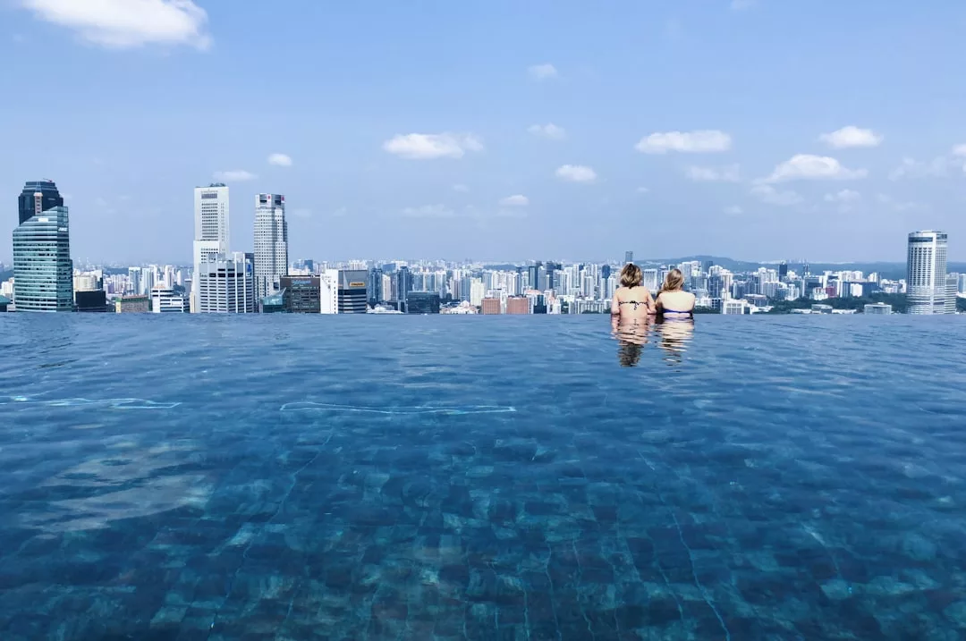 Two people stand at the edge of an infinity pool overlooking a city skyline, embracing a luxe lifestyle under a blue sky with scattered clouds.