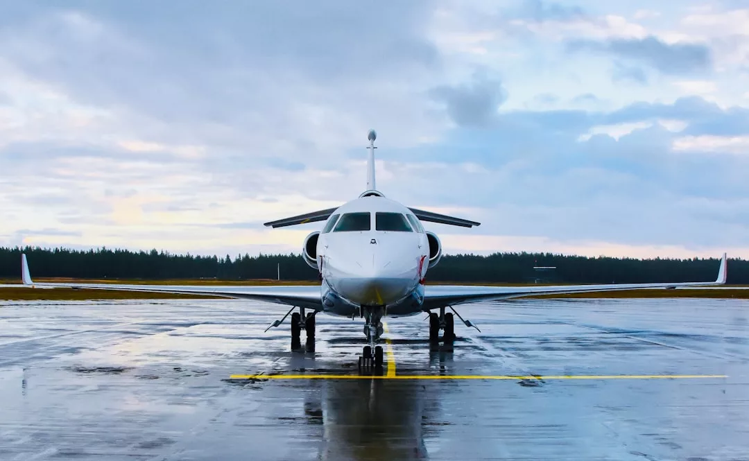 A white private jet is parked on a wet runway, reflecting a lifestyle of luxury with trees and a cloudy sky in the background.